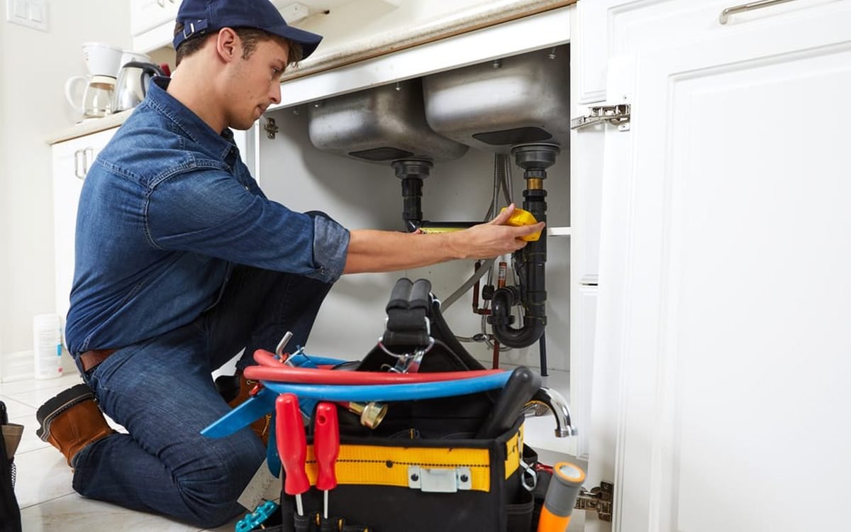 Plumber repairing pipes under a kitchen sink with a full tool belt